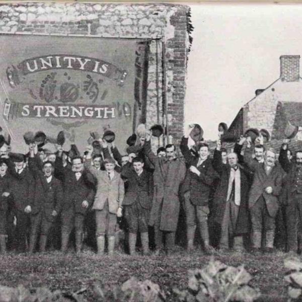 An old, black and white picture of a group of people stood in front of a banner saying "Unity is Strength"